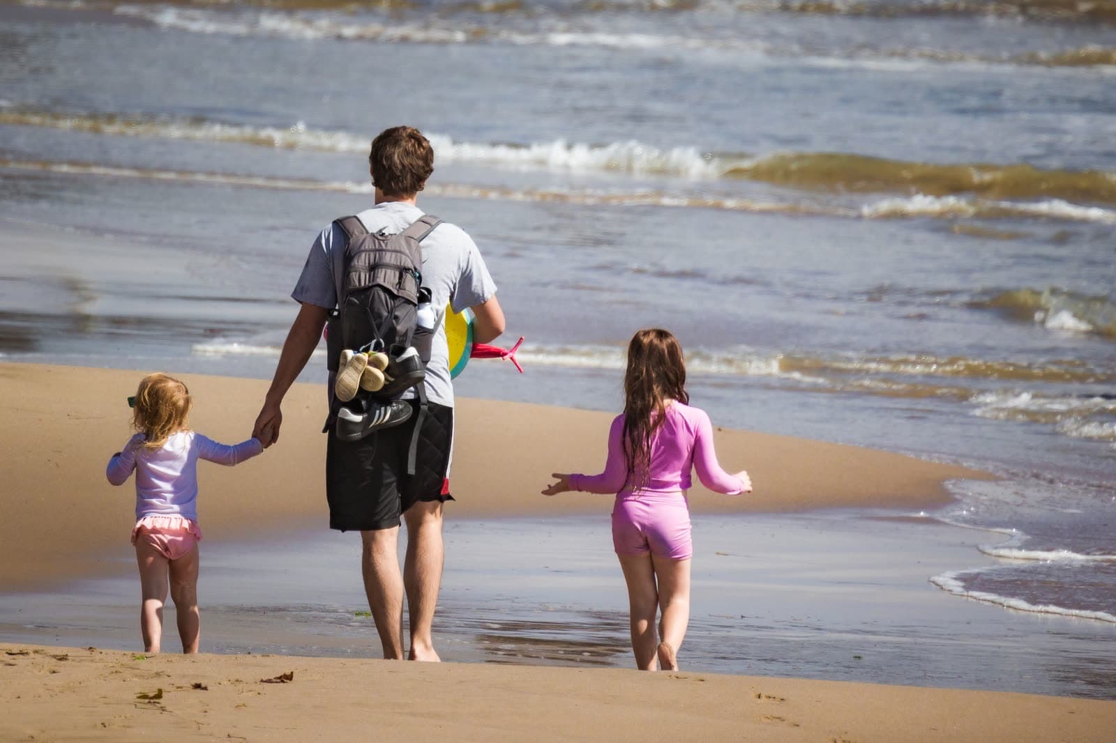 A father and two young girls, seen from behind, walking hand in hand along the shoreline